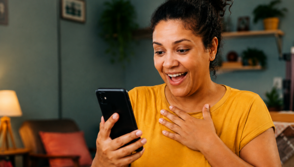 Mulher sorri ao olhar o celular em casa, representando o uso de aplicativos para ganhar dinheiro e fazer renda extra pelo celular.