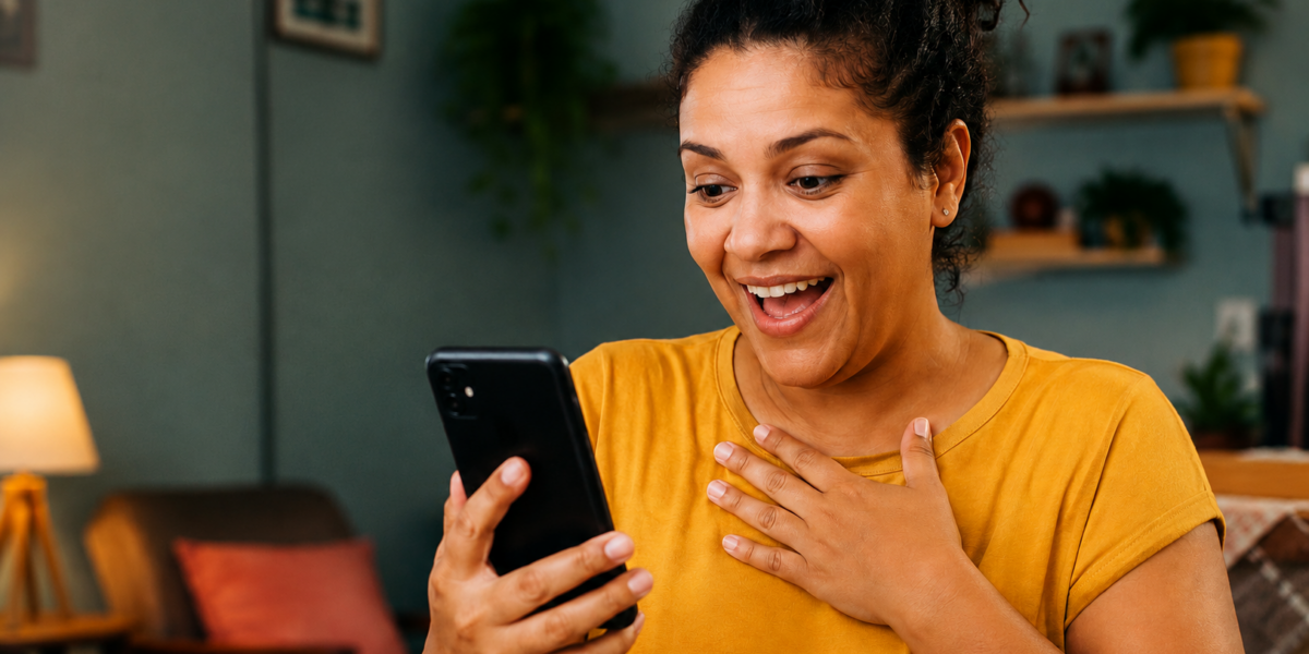 Mulher sorri ao olhar o celular em casa, representando o uso de aplicativos para ganhar dinheiro e fazer renda extra pelo celular.