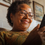 Mulher sorrindo enquanto acessa o app da Tele Sena pelo celular em casa.