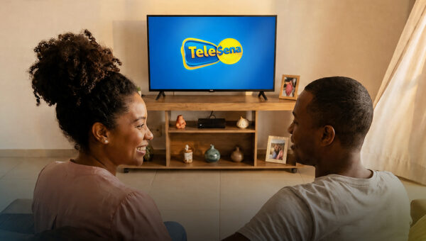 Couple sitting on a couch watching a TV that displays the Tele Sena logo.