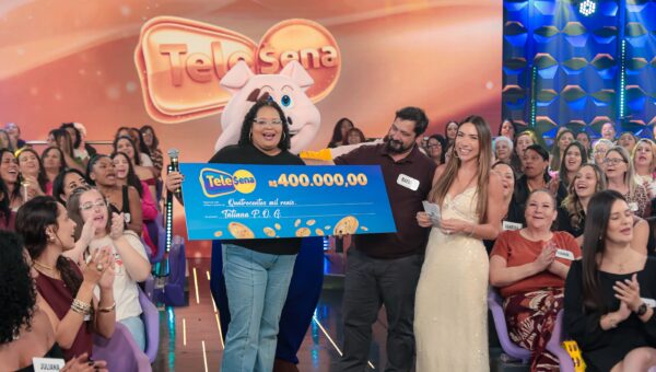 Ganhadora da Tele Sena recebe prêmio de R$ 400 mil no palco do SBT, com plateia comemorando.