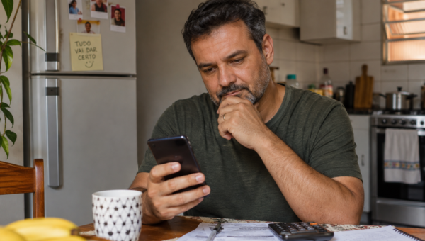 Homem olhando o celular enquanto organiza contas e despesas sobre a mesa, representando o uso do Pix parcelado.