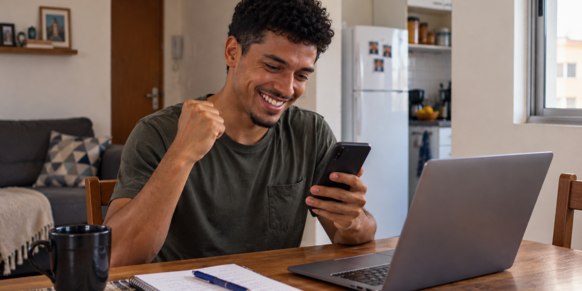 Homem jovem sorrindo ao usar o celular ao lado de um notebook, representando formas de ganhar dinheiro rápido e renda extra.